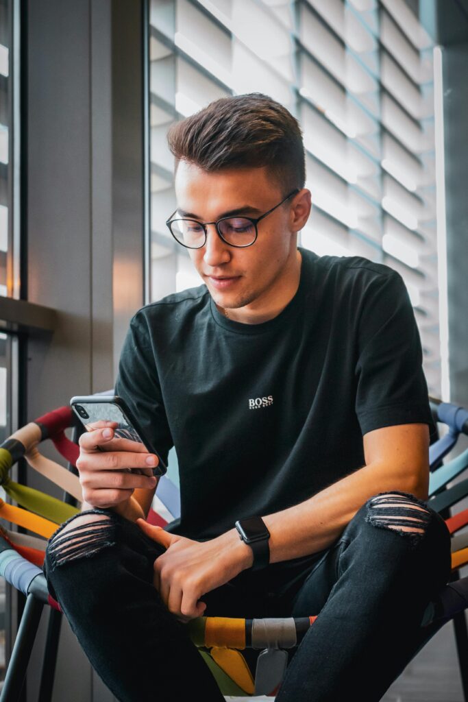 A fashionable young man with glasses using a smartphone indoors, sitting comfortably.
